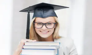 Uśmiechnięta studentka w birecie i okularach trzyma stos książek, zastanawiając się, ile trwają studia podyplomowe.