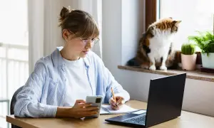 Kobieta w okularach pracuje zdalnie okazjonalnie, notując coś w zeszycie i patrząc na telefon. Kot obserwuje świat za oknem.