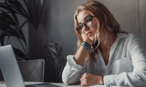 Kobieta w okularach opiera głowę na dłoni, patrząc na laptopa. Brak jej motywacji do pracy, ale wierzy, że cel jest blisko.