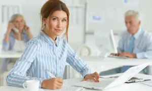 Kobieta na kursie księgowości pracuje przy laptopie, notując coś na papierze. W tle widać innych pracowników biurowych.