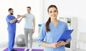 Uśmiechnięta asystent medyczny z tablicą w ręku, w tle fizjoterapeuta pracuje z pacjentem.