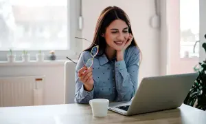 Uśmiechnięta kobieta z okularami w dłoni pracuje przy laptopie. To może być jej dzień w dziale **human resources**.