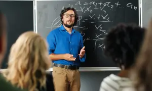 Nauczyciel w niebieskiej koszuli tłumaczy matematykę przy tablicy. Czy nauczyciel jest funkcjonariuszem publicznym? To pytanie nurtuje wielu.