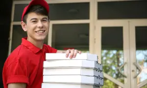 Uśmiechnięty młodociany dostawca pizzy w czerwonej koszulce i czapce trzyma stos pudełek z pizzą przed drzwiami.