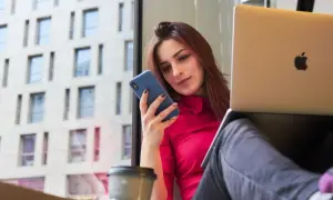 Kobieta w czerwonej bluzce korzysta z telefonu i laptopa, pracując w domu.