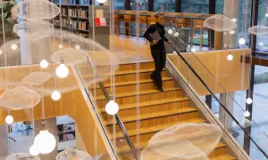 Kobieta schodzi po drewnianych schodach w nowoczesnej bibliotece, niosąc laptopa. Zastanawia się, ile trwają studia.