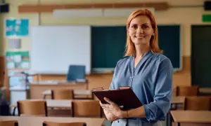 Uśmiechnięta nauczycielka z teczką w ręku, w tle klasa szkolna. Legitymacja nauczyciela to symbol jej pracy i zaangażowania.