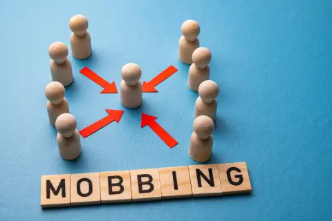Drewniane pionki otaczają jednego, a czerwone strzałki wskazują na niego. Słowo "MOBBING" ułożone z klocków. Jak udowodnić mobbing w pracy?