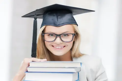 Uśmiechnięta studentka w birecie i okularach trzyma stos książek, zastanawiając się, ile trwają studia podyplomowe.
