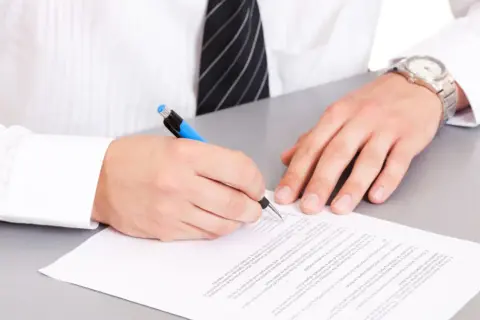 Mężczyzna w białej koszuli i krawacie podpisuje dokument. Na papierze widoczna jest klauzula CV po angielsku.