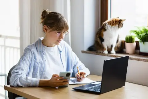Kobieta w okularach pracuje zdalnie okazjonalnie, notując coś w zeszycie i patrząc na telefon. Kot obserwuje świat za oknem.