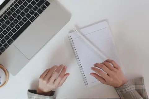 Ręce piszą notatki do CV po niemiecku przy laptopie, obok filiżanka kawy.