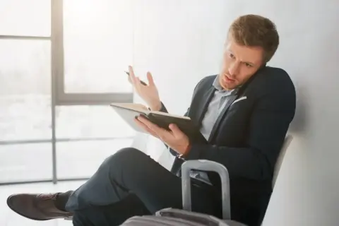 Mężczyzna w garniturze siedzi z notatnikiem i telefonem przy uchu, czekając na delegację.