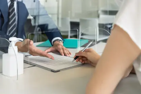 Pracownik samorządowy w garniturze wskazuje na dokument, który podpisuje klientka. Obok stoi płyn do dezynfekcji.