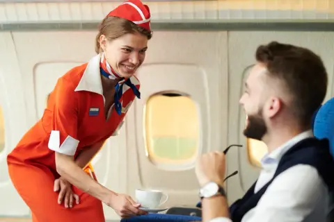 Uśmiechnięta stewardessa w pomarańczowym stroju podaje kawę pasażerowi w samolocie.