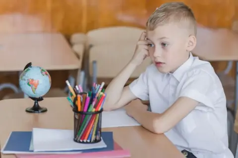 Chłopiec zamyślony przy biurku, na którym leży globus i przybory. Obowiązek szkolny bywa trudny.