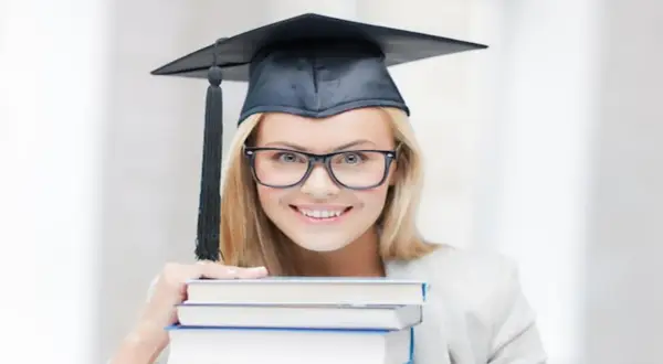 Uśmiechnięta studentka w birecie i okularach trzyma stos książek, zastanawiając się, ile trwają studia podyplomowe.