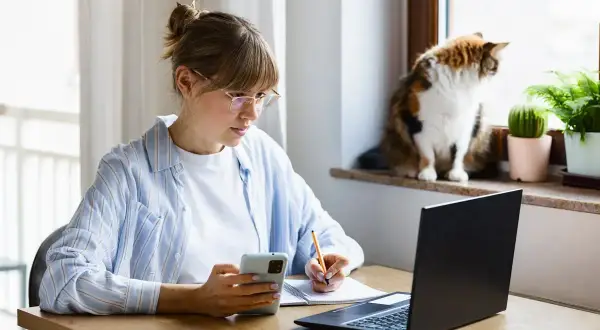 Kobieta w okularach pracuje zdalnie okazjonalnie, notując coś w zeszycie i patrząc na telefon. Kot obserwuje świat za oknem.