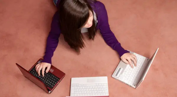 Kobieta leży na dywanie i pisze na trzech laptopach.