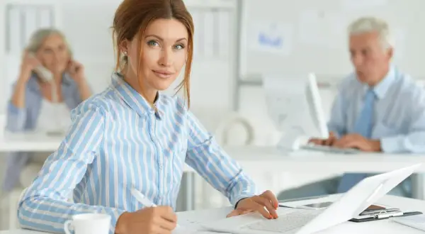 Kobieta na kursie księgowości pracuje przy laptopie, notując coś na papierze. W tle widać innych pracowników biurowych.