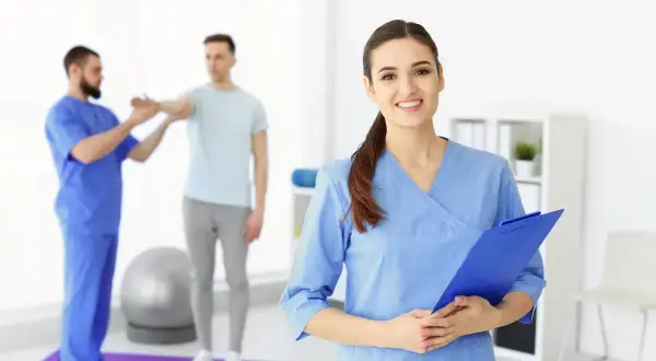 Uśmiechnięta asystent medyczny z tablicą w ręku, w tle fizjoterapeuta pracuje z pacjentem.