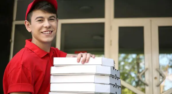 Uśmiechnięty młodociany dostawca pizzy w czerwonej koszulce i czapce trzyma stos pudełek z pizzą przed drzwiami.
