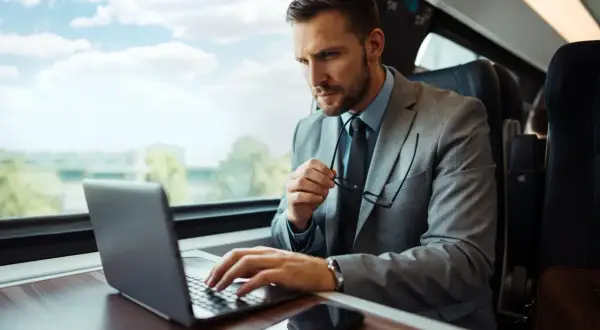 Biznesmen w garniturze pracuje na laptopie w pociągu, trzymając okulary.