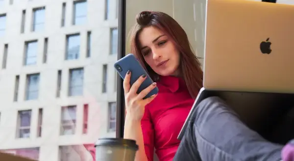 Kobieta w czerwonej bluzce korzysta z telefonu i laptopa, pracując w domu.