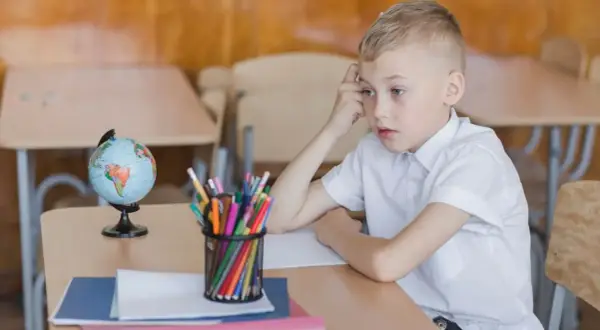 Chłopiec zamyślony przy biurku, na którym leży globus i przybory. Obowiązek szkolny bywa trudny.