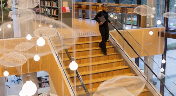 Kobieta schodzi po drewnianych schodach w nowoczesnej bibliotece, niosąc laptopa. Zastanawia się, ile trwają studia.