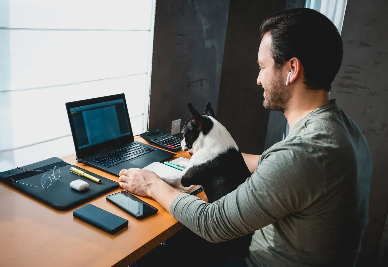 Mężczyzna z psem pracuje zdalnie bez doświadczenia. Na biurku laptop, telefon, okulary i kalkulator.