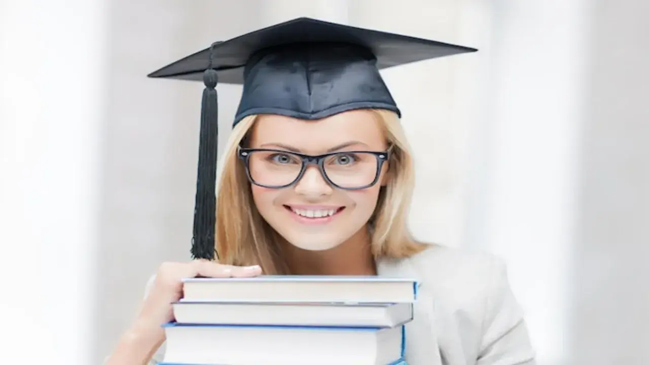 Uśmiechnięta studentka w birecie i okularach trzyma stos książek, zastanawiając się, ile trwają studia podyplomowe.