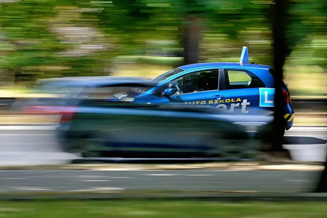 Niebieski samochód z napisem "AUTO SZKOŁA" i literą "L" na drzwiach, w ruchu. Idealny do kursu na instruktora nauki jazdy.