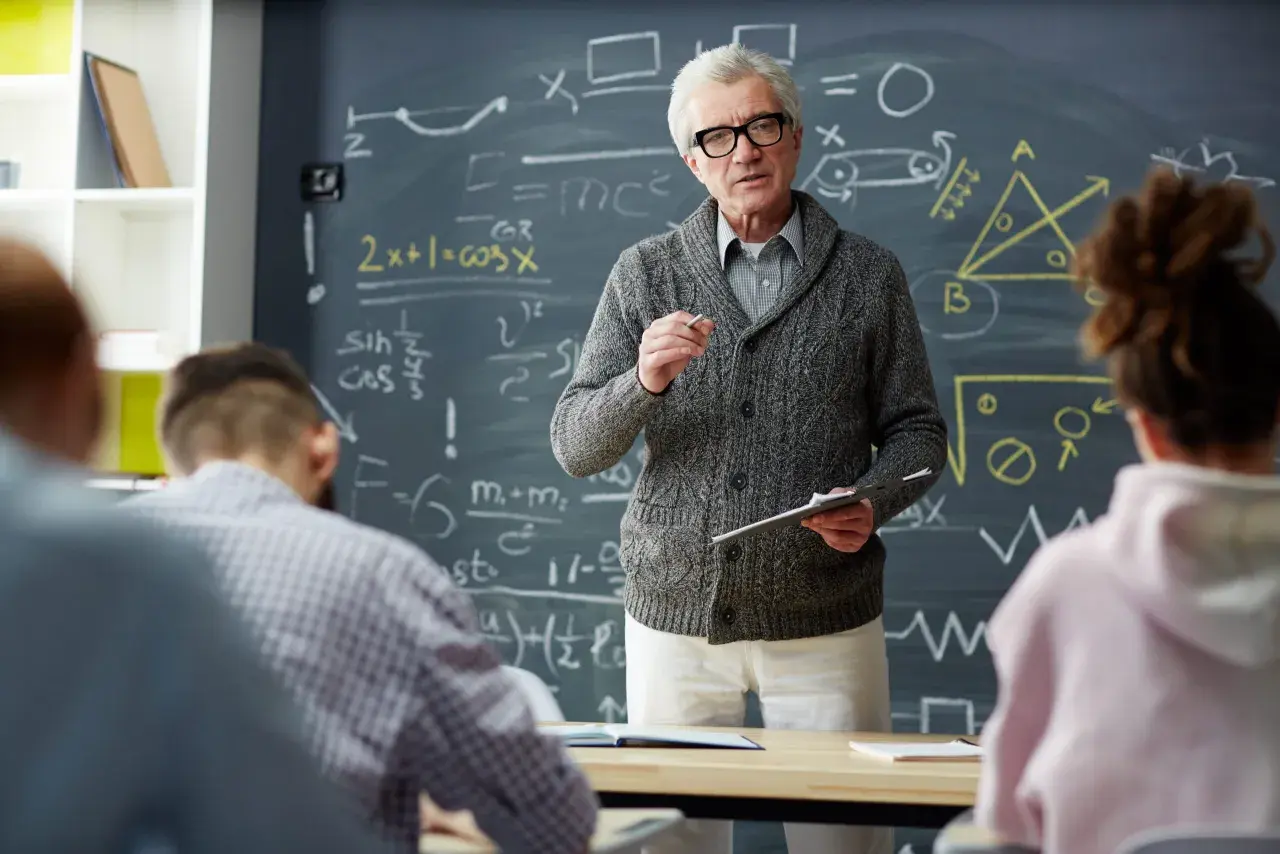 Nauczyciel z tablicą pełną wzorów matematycznych wyjaśnia coś uczniom. Zastanawiasz się, ile zarabia nauczyciel początkujący?