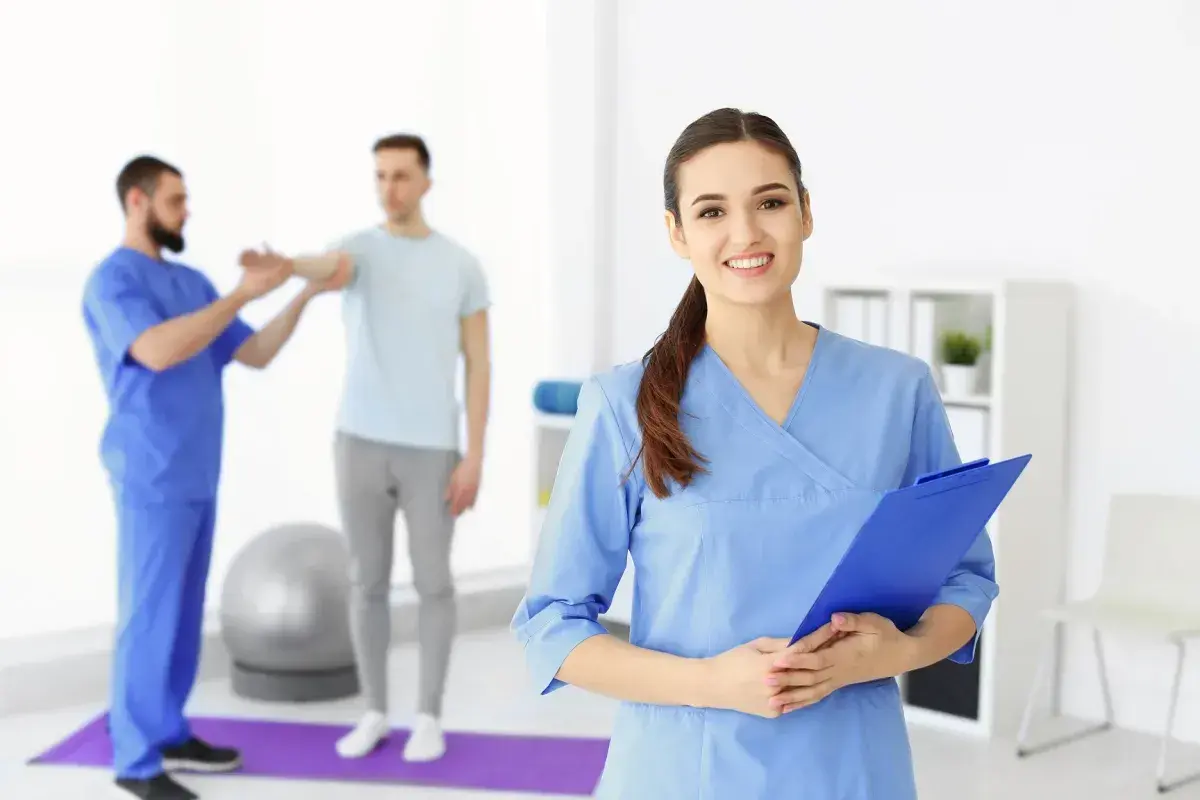 Uśmiechnięta asystent medyczny z tablicą w ręku, w tle fizjoterapeuta pracuje z pacjentem.