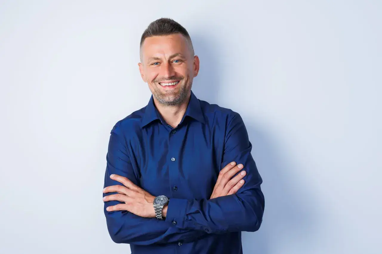Uśmiechnięty mężczyzna w granatowej koszuli, z rękami skrzyżowanymi na piersi. Idealne zdjęcie do CV zrobione telefonem.