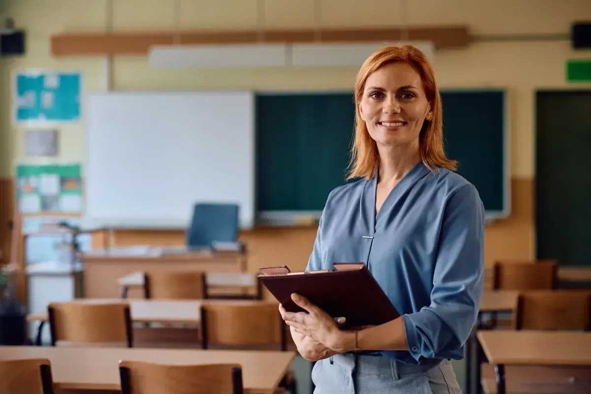 Uśmiechnięta nauczycielka z teczką w ręku, w tle klasa szkolna. Legitymacja nauczyciela to symbol jej pracy i zaangażowania.