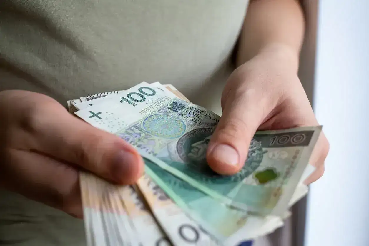 Dłonie trzymają stos banknotów 100 zł. To kwota, która dla wielu jest znacząca, ale w kontekście dużych inwestycji może być traktowana jako de minimis.