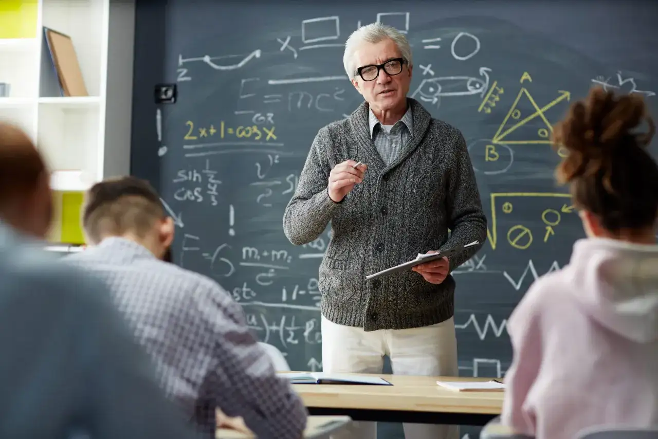 Nauczyciel z tablicą pełną wzorów matematycznych wyjaśnia coś uczniom. Zastanawiasz się, ile zarabia nauczyciel początkujący?