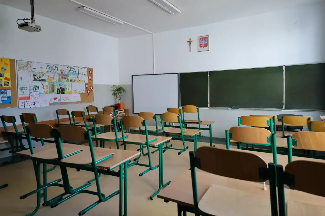 Pusta klasa szkolna z rzędami ławek i krzeseł. Na ścianie wiszą rysunki dzieci, tablica i godło Polski. Niska frekwencja w szkole?