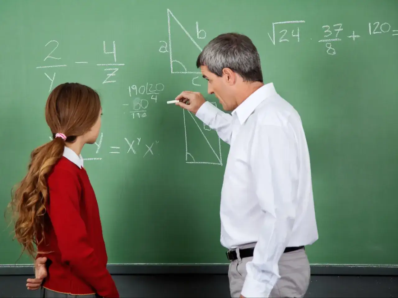 Nauczyciel matematyki pisze kredą na tablicy, obok stoi uczennica.