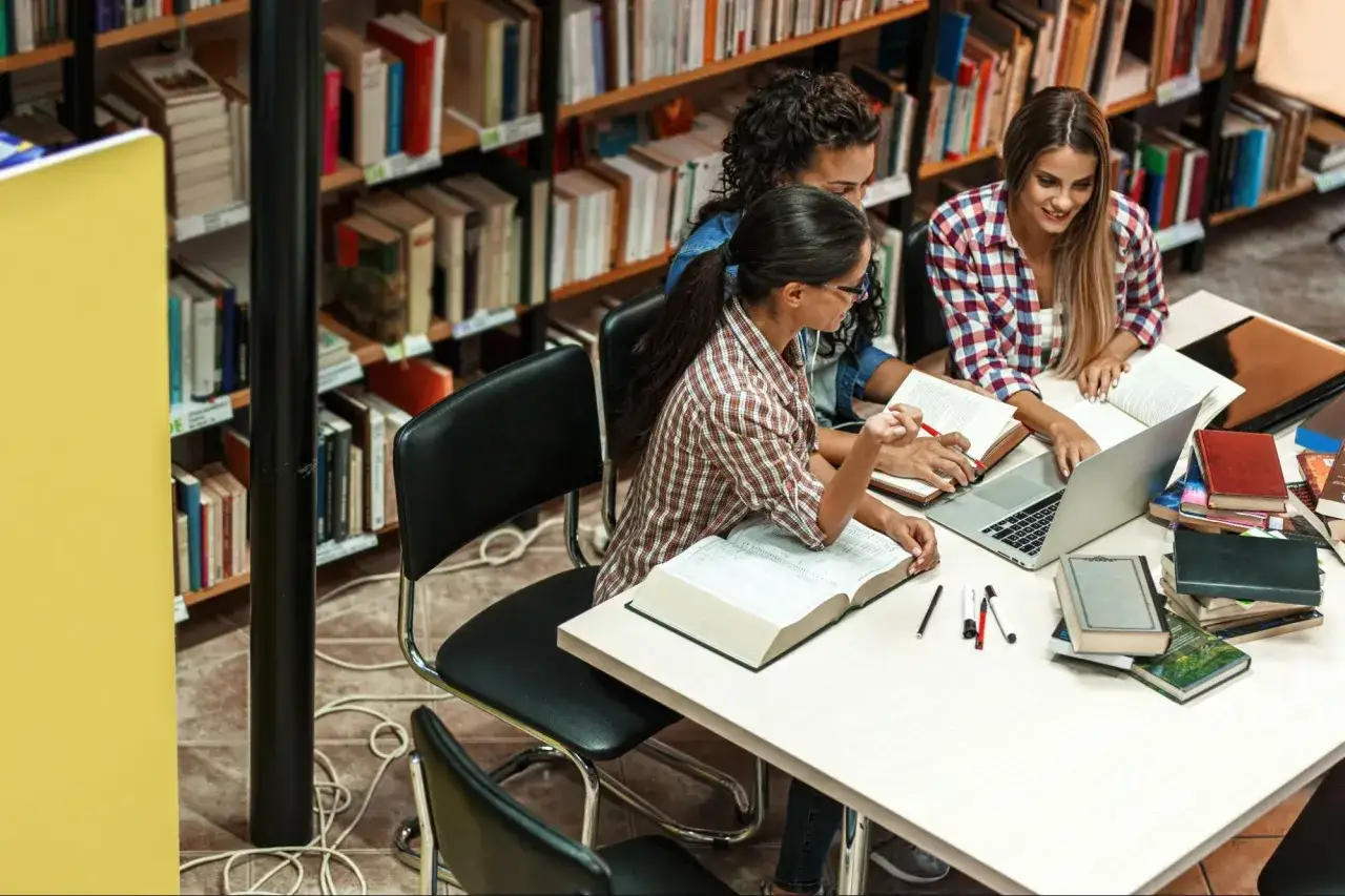 Trzy studentki uczące się w bibliotece, otoczone książkami, zdobywają wykształcenie wyższe.
