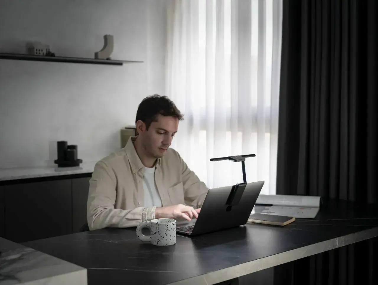 Mężczyzna pracuje przy laptopie, czerpiąc motywację do pracy z otoczenia. Na biurku kubek i notatnik.