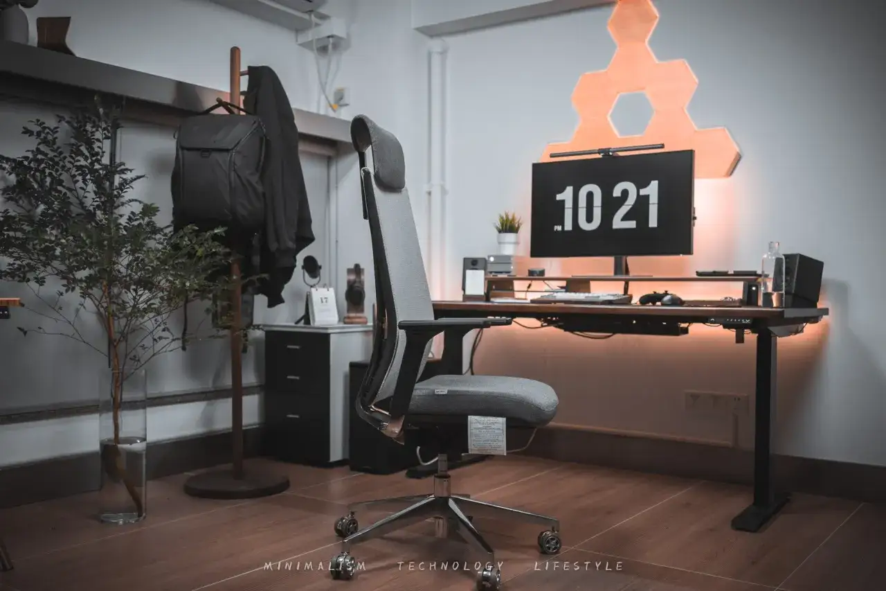 Nowoczesne stanowisko do pracy w domu: ergonomiczne krzesło, biurko z regulacją wysokości, monitor z zegarem i dekoracyjne oświetlenie.