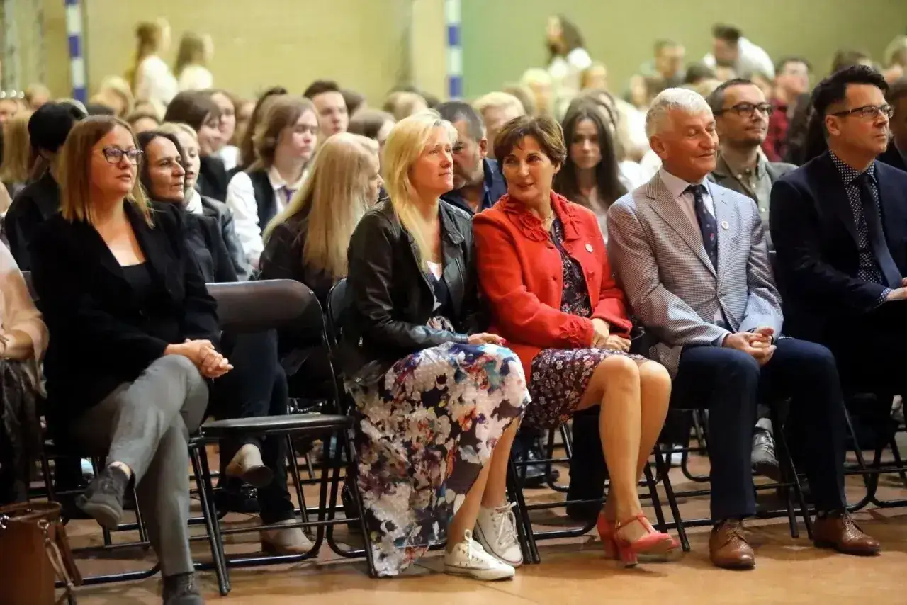 Uczestnicy siedzą na krzesłach, słuchając uważnie. Wśród nich widać osoby, które mogą być na radzie pedagogicznej, w tym kobiety w eleganckich strojach i mężczyzn w garniturach.