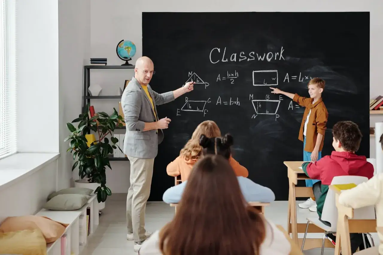 Nauczyciel i uczeń wyjaśniają wzory geometryczne na tablicy. To zajęcia dydaktyczno wyrównawcze z matematyki.