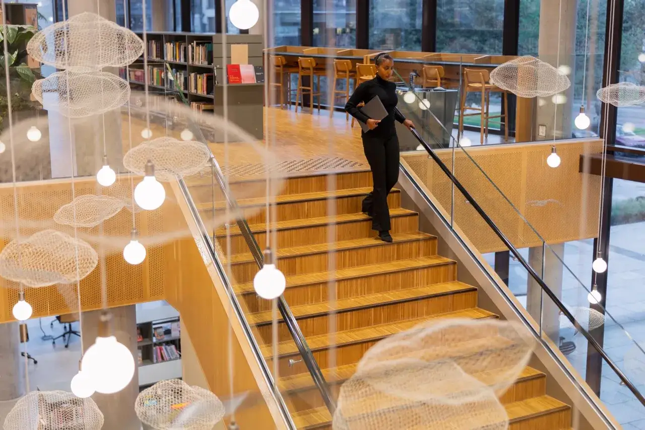 Kobieta z laptopem schodzi po drewnianych schodach w nowoczesnej bibliotece. Zastanawia się, ile trwają studia, gdy mijają ją wiszące dekoracje w kształcie chmur.