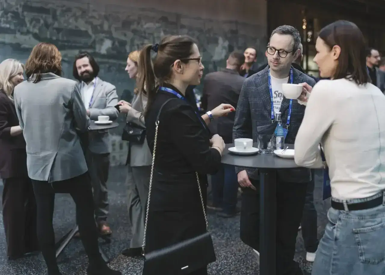 Ludzie na konferencji rozmawiają przy stolikach. To świetny moment, by dowiedzieć się, jak zacząć rozmowę i nawiązać nowe kontakty.