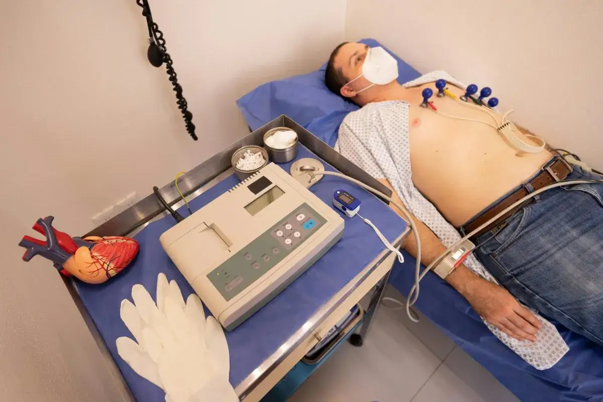 Pacjent z maską na twarzy leży podłączony do aparatury EKG. Obok leży model serca, rękawiczki i pulsoksymetr. To praca asystenta medycznego.