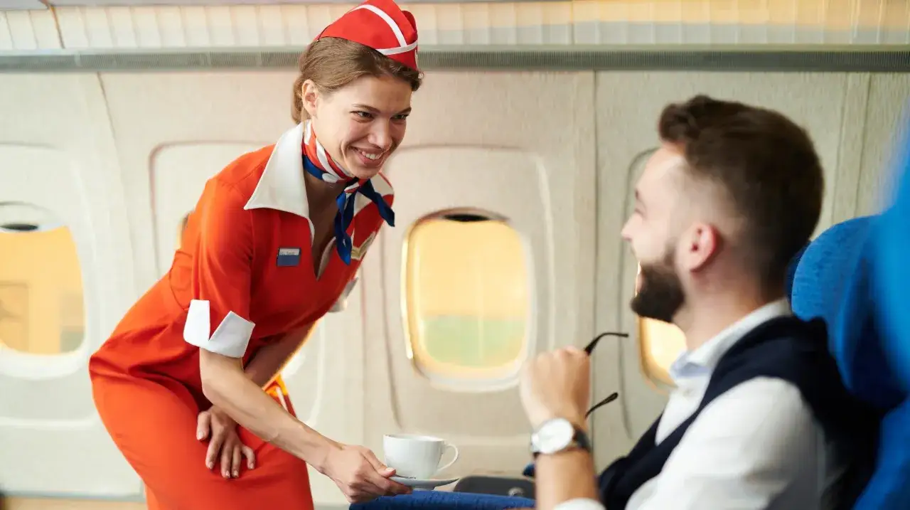 Uśmiechnięta stewardessa w pomarańczowym stroju podaje kawę pasażerowi w samolocie.